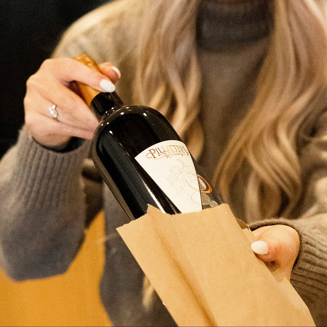 A person in a gray sweater holds a dark wine bottle partially tucked into a brown paper bag. The bottle label appears to read Pillitteri Estates Winery, and the scene is photographed in a real indoor setting with warm lighting.