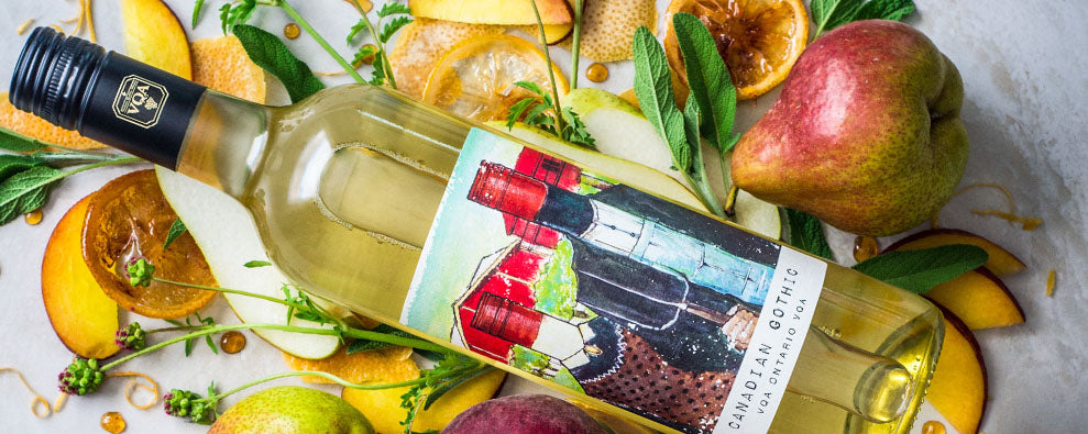 A clear bottle with a black screw cap filled with white wine. The bottle is laying down and is surrounded by fruits including pears, oranges and mango. 