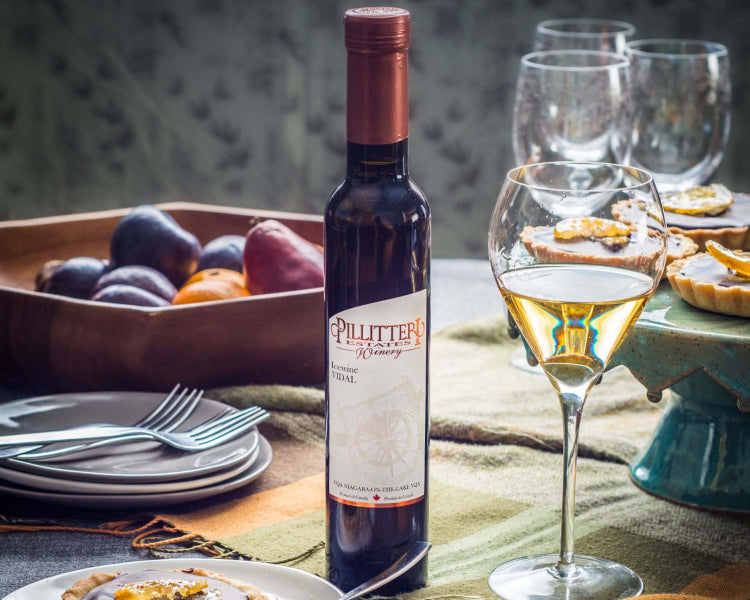 A bottle of Pillitteri Vidal Icewine stands on a styled dining table beside a filled wine glass and plates of food, with wine glasses in the background.