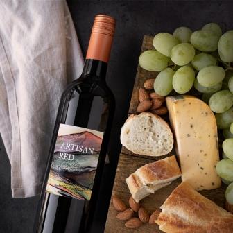 A bottle of Artisan Red wine positioned next to a selection of cheeses, bread, and grapes on a wooden board, with a napkin to the side.